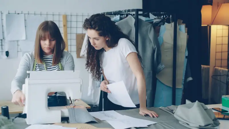 Two designers working at a sewing machine in a studio with garment racks