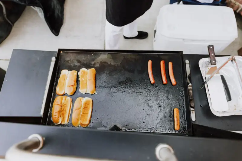 Hot dogs and buns cooking on a portable griddle beside a cooler