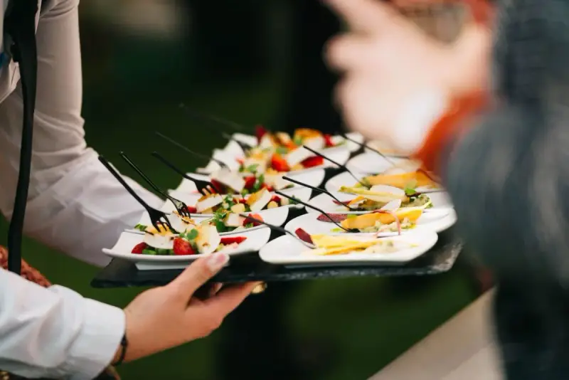 A server carries a tray of individually plated cheese, fruit and vegetable appetizers with small forks.