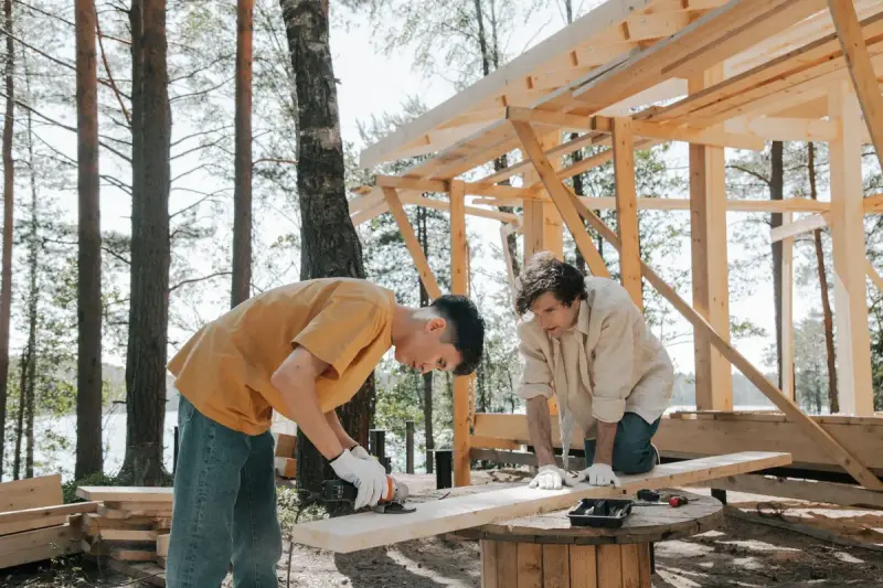 Two carpenters cutting and checking a board under a timber frame among trees
