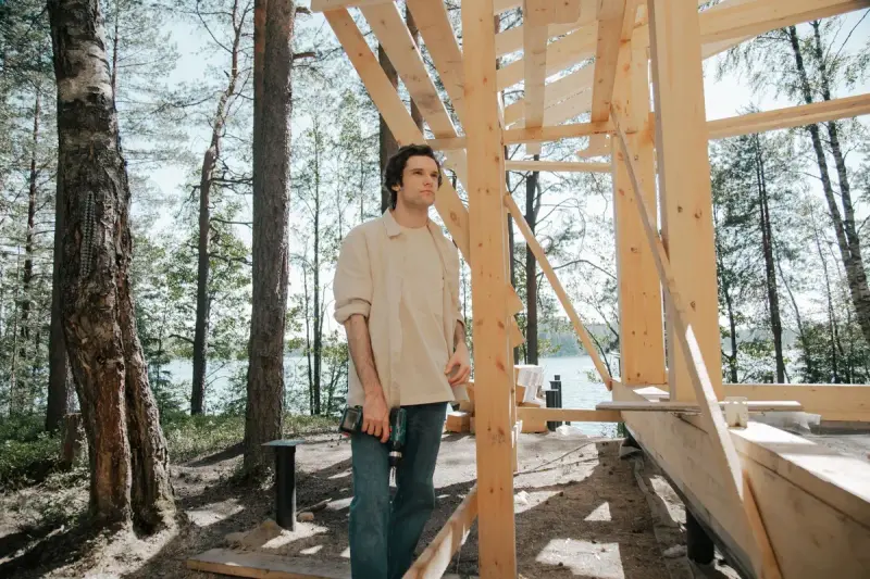 Carpenter holding a cordless drill beside a timber house frame by a lake
