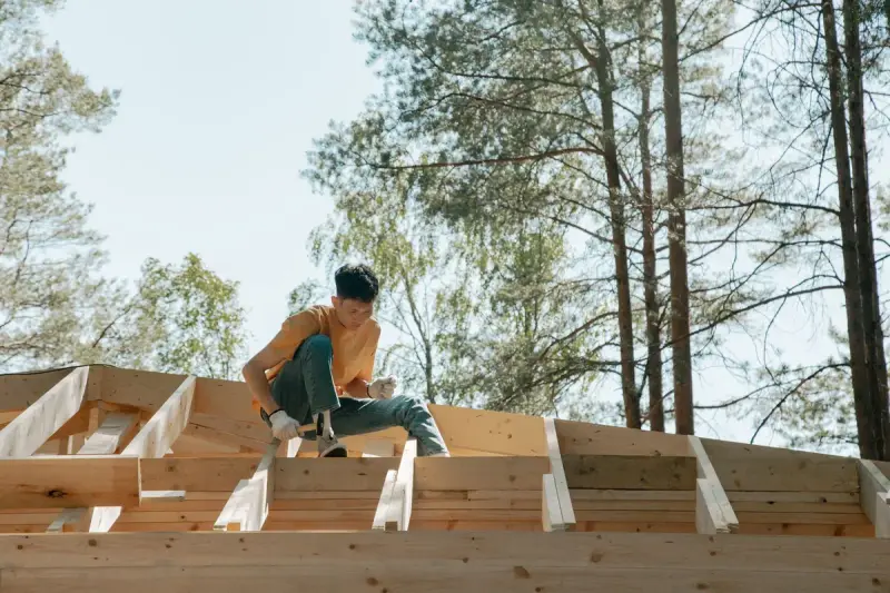 Carpenter seated on new wooden roof trusses with tall pines behind