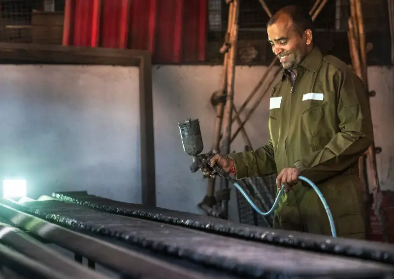 Smiling worker in coveralls spraying a long metal part with a spray gun