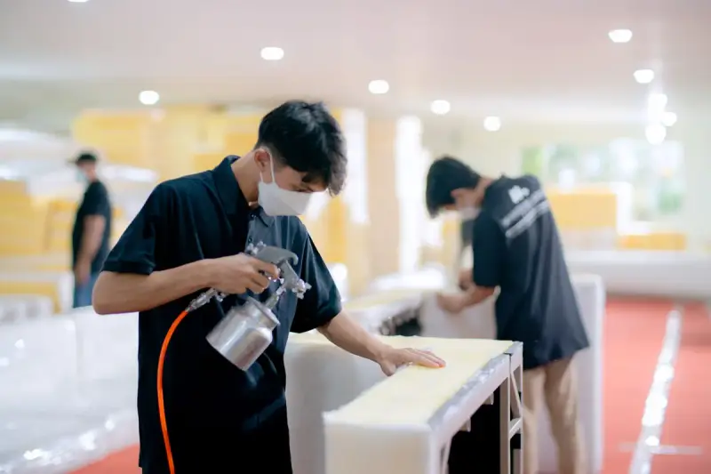 Masked workers spray-finishing white furniture panels in a bright factory finishing area