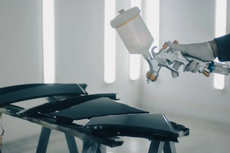 Gloved hand aiming a gravity-feed spray gun at dark panels on stands in a white booth