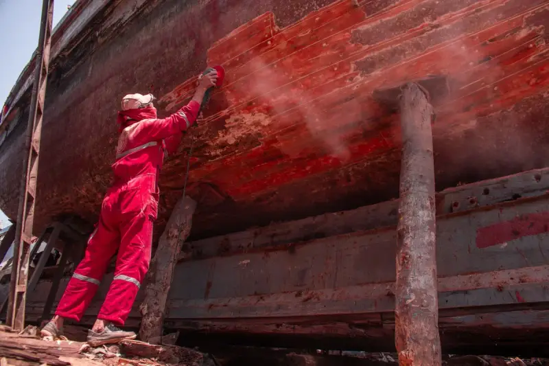 Worker in red coveralls and mask sanding the red hull of a ship in a shipyard