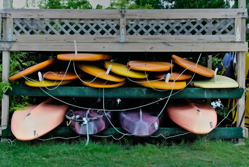 Stacked paddleboards and kayaks on an outdoor wooden rack beside a lattice fence