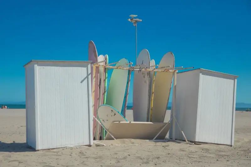 Surfboards standing in a wooden rack between white beach cabins under a deep blue sky