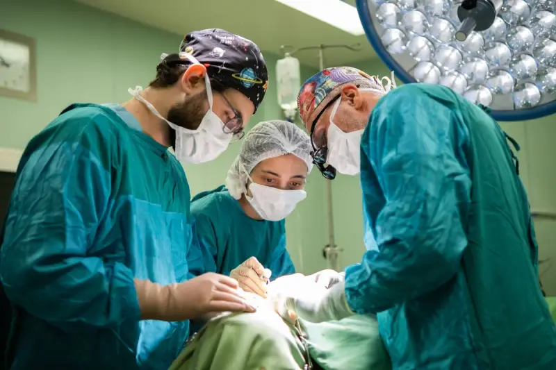 Three surgeons in teal gowns and caps, one woman, operate together under a surgical light