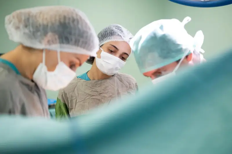 A woman surgeon in cap and mask watches closely beside two colleagues in the operating room