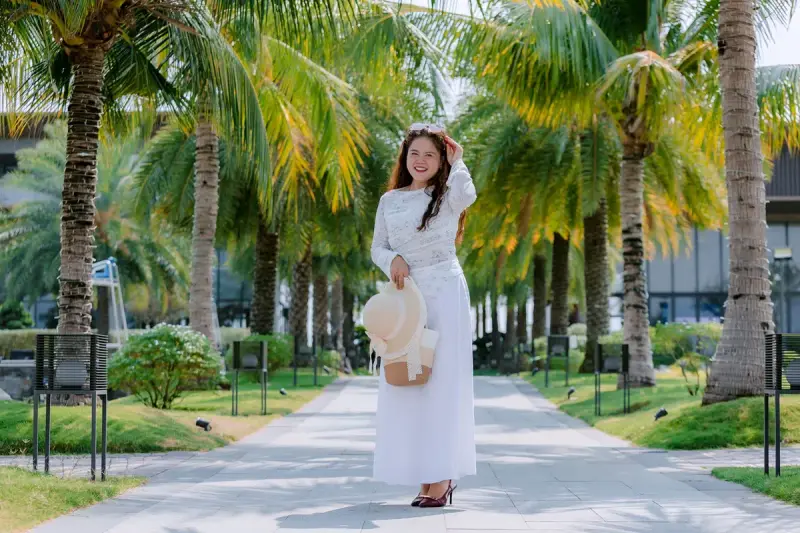 Woman in a long white dress holding a straw hat on a path under tall palm trees