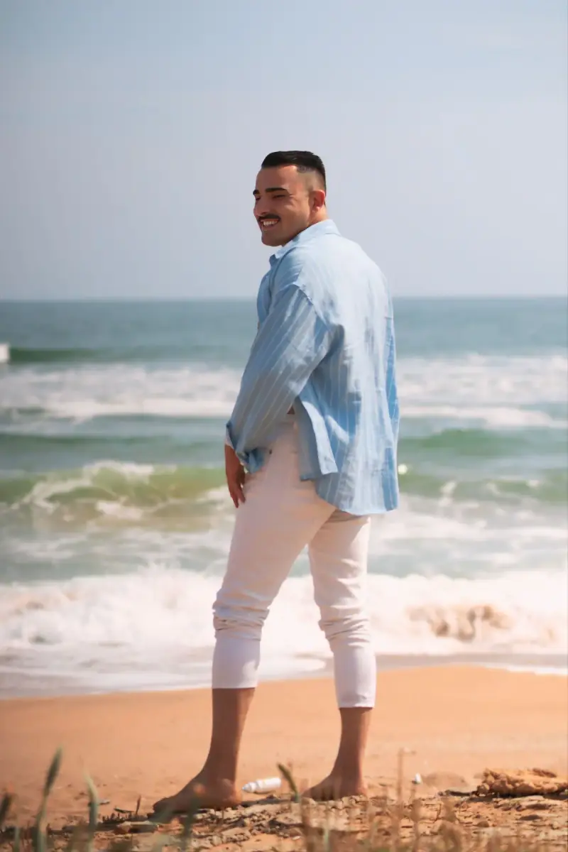 Man in a pale blue linen shirt and white trousers barefoot at the surf line