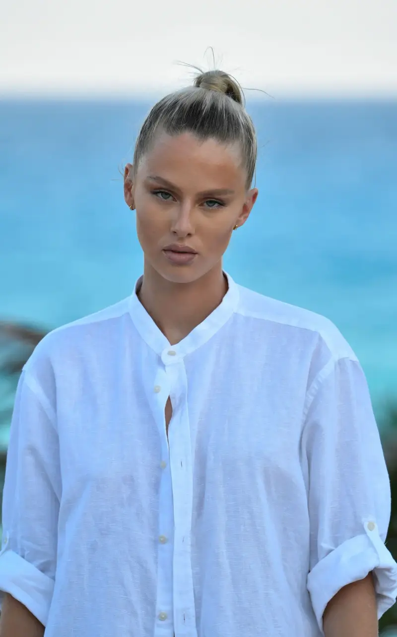 Woman in a white band-collar linen shirt with turquoise sea behind her
