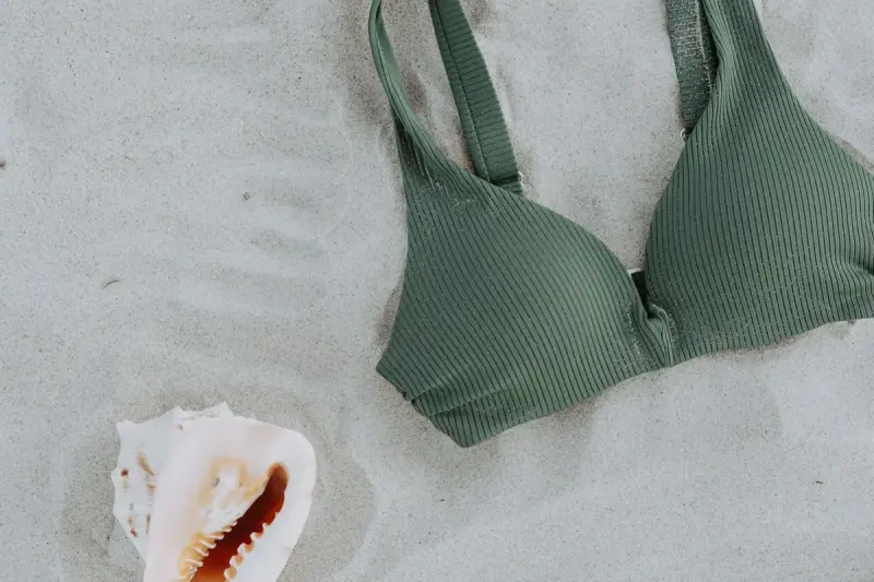 A green bikini top and a seashell laid flat on sand