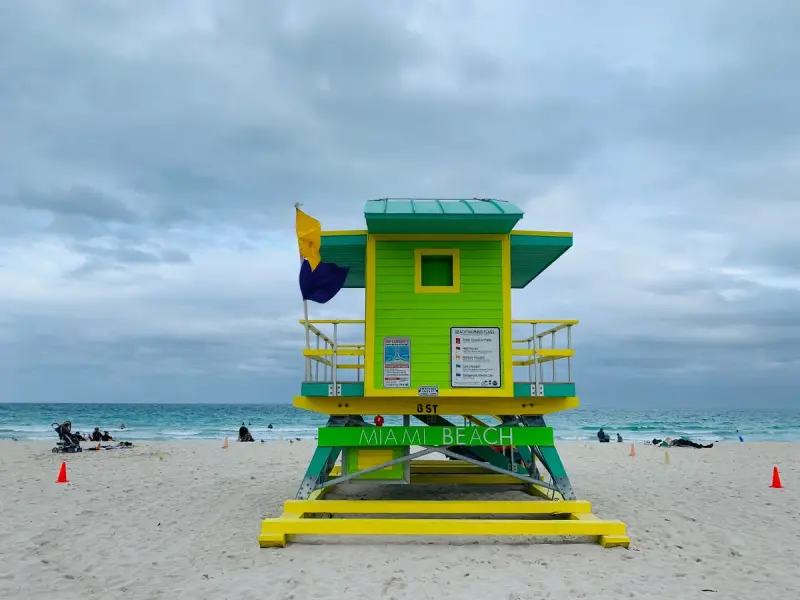 A yellow and green lifeguard house marked Miami Beach on the sand by the sea