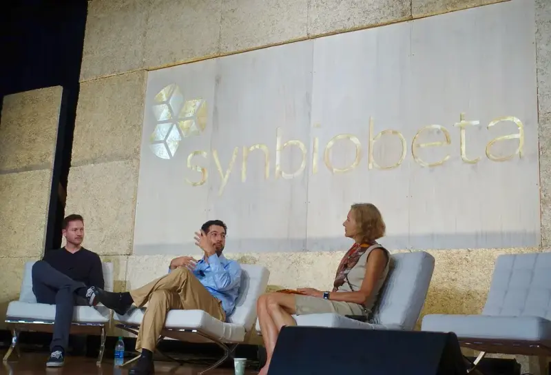 Three panelists in white chairs on stage under a large SynBioBeta sign