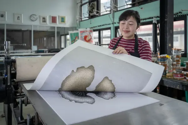 A printmaker lifts a printed sheet beside a press in a printmaking workshop. Paper- photograph.