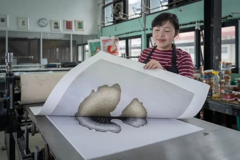 A printmaker lifts a printed sheet beside a press in a printmaking workshop. Paper- photograph.