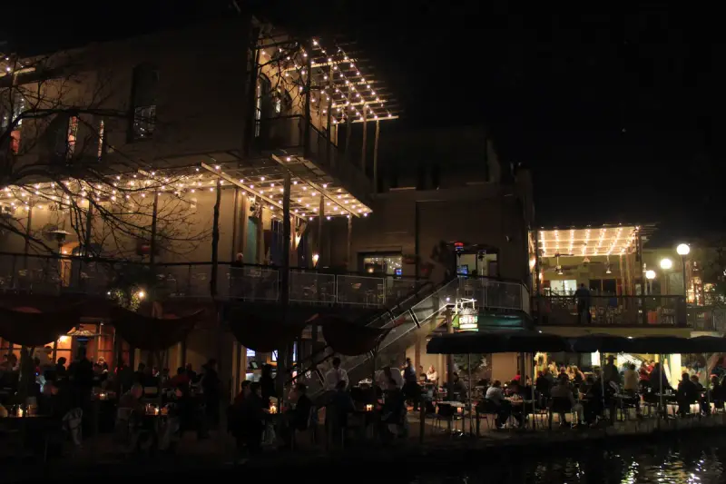 Diners at candlelit tables on a lit restaurant terrace along the San Antonio River Walk at night