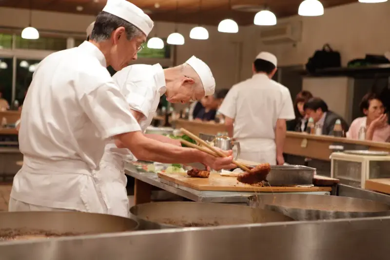 Two chefs in white uniforms plating fried cutlets at an open kitchen counter