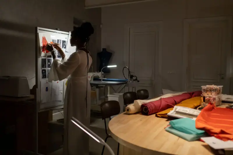 Designer pinning work to a lit board in a studio stacked with fabric