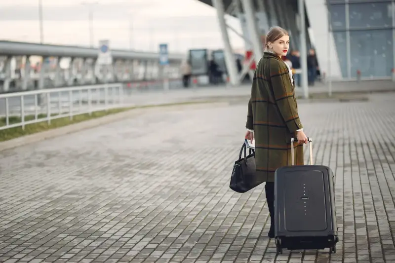 Woman in a plaid coat with a black carry-on and tote walking outside an airport terminal
