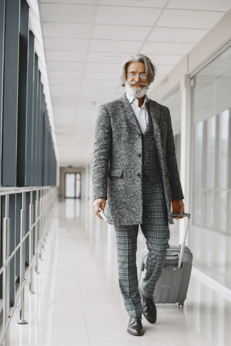 Silver-haired man in a herringbone coat and check suit rolling a carry-on down a bright corridor