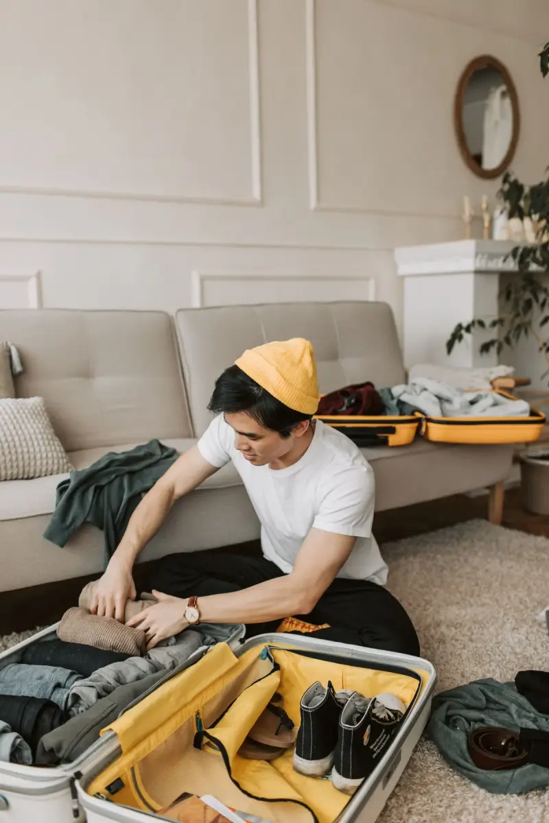 Man in a mustard beanie packing clothes into an open yellow-lined suitcase on the floor