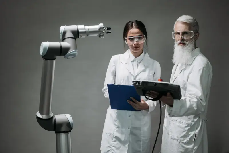 Woman and man in lab coats and safety glasses reviewing data beside a collaborative robot arm