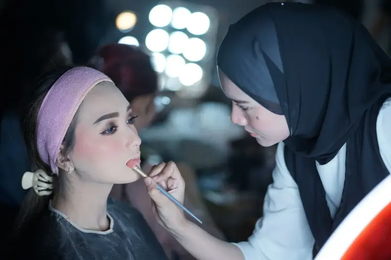 Makeup artist in a hijab lining a model's lips under vanity bulbs