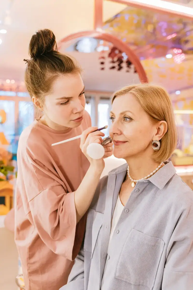 Young makeup artist applying color to an older woman in a pink beauty studio