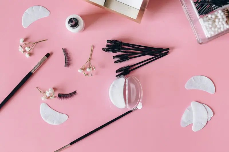 Lash strips, spoolies, eye pads and brushes arranged on a pink surface