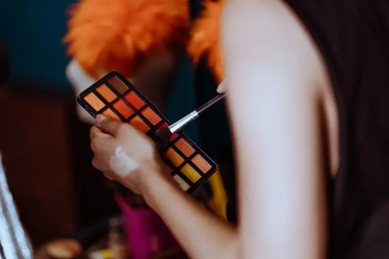 Artist holding an orange eyeshadow palette and brush in a dressing room