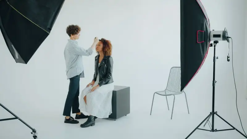 Makeup artist touching up a seated subject in a white photo studio with softboxes