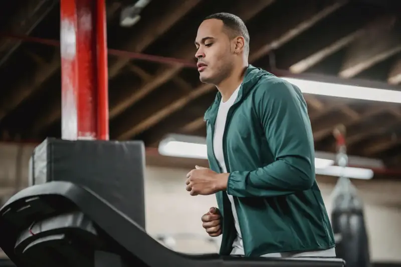 Man in a green running jacket jogging on a treadmill in an industrial-style gym