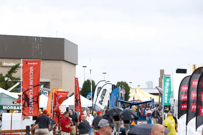 Visitors with umbrellas walking an outdoor aisle of exhibitor banners and equipment