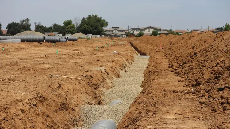 Long trenches and stacked pipe across a suburban underground construction site