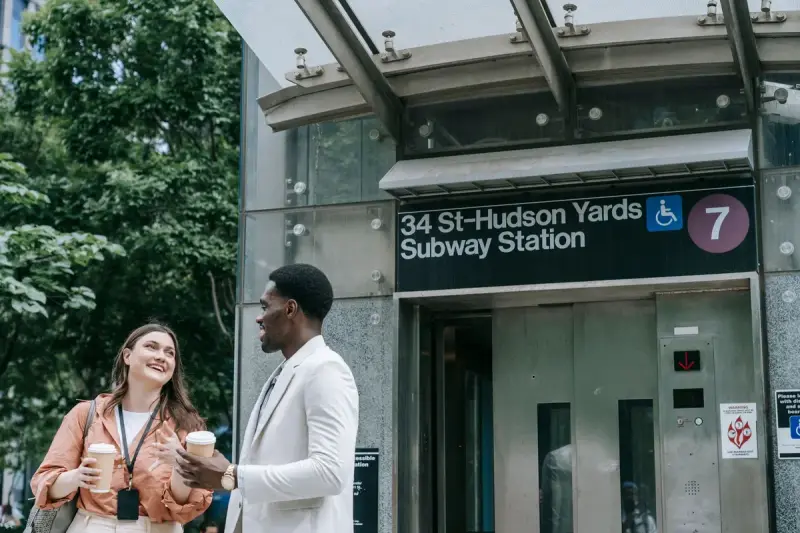 Two colleagues wearing ID badges talk over coffee outside the 34 St–Hudson Yards subway station