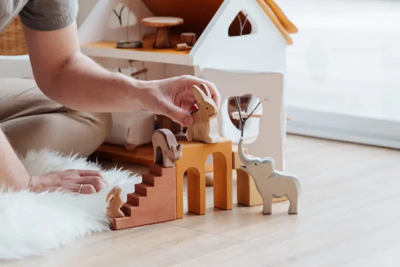 An adult's hands arranging wooden animal toys beside a white wooden toy house on the floor