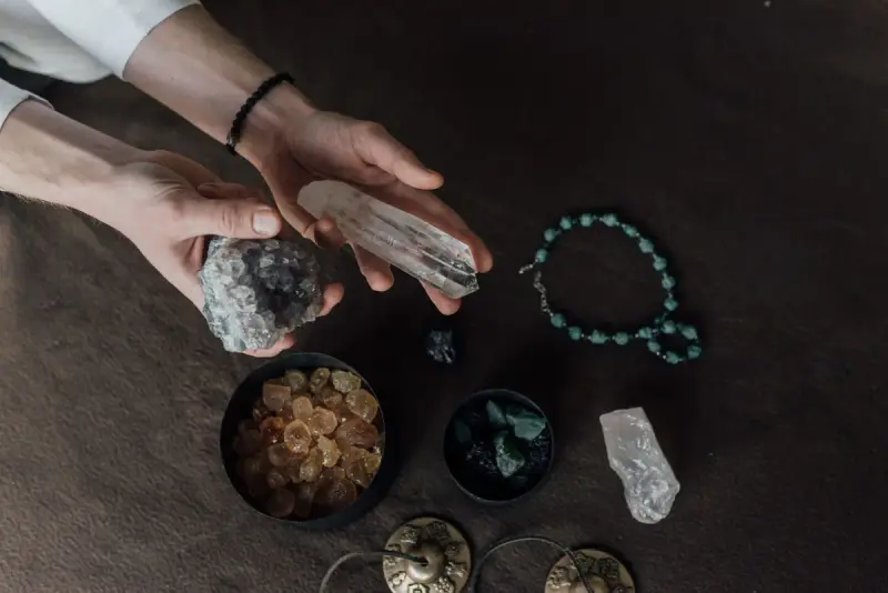 Hands holding a raw crystal over bowls of loose stones and a beaded bracelet on a dark table