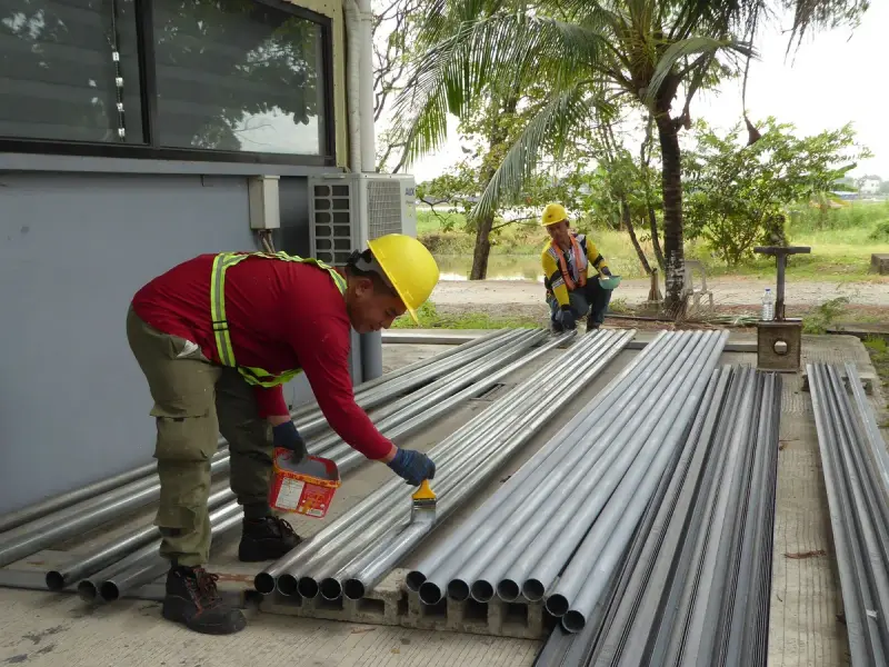 Worker in hard hat and hi-vis vest coating a row of metal pipes outdoors