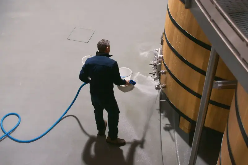 Cellar worker hosing down the floor beside large oak vats in a winery