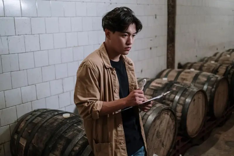 Cellar worker with a clipboard checking rows of oak barrels in a white-tiled cellar