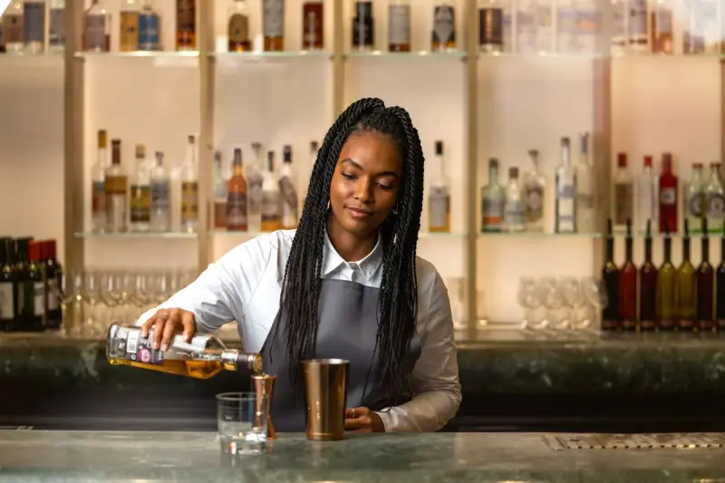 Bartender with braided hair pouring spirits into a copper mixing cup at a bar