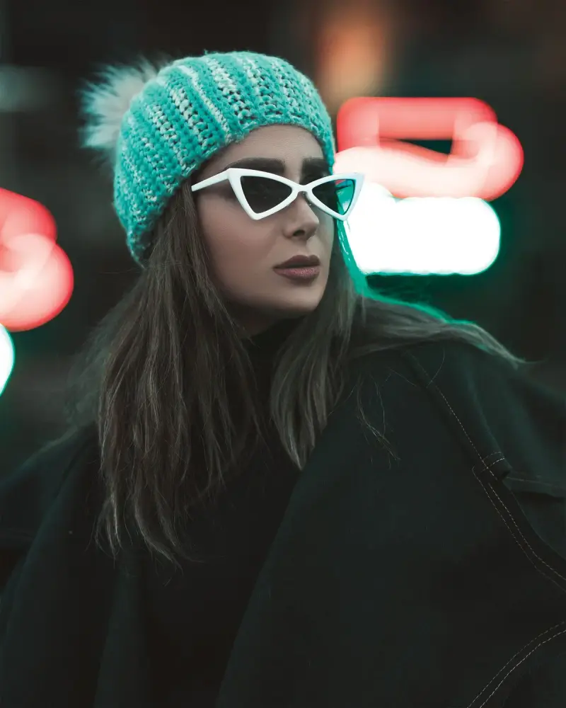 Woman in white angular cat-eye sunglasses and a knit beanie against neon lights