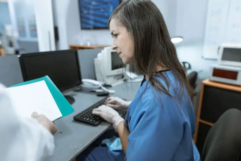 Nurse in blue scrubs and gloves types at a workstation while a colleague holds a folder
