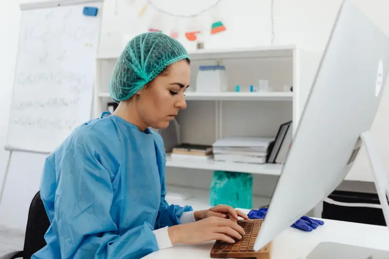 Woman in a blue gown and hair cover types on a keyboard in front of a large monitor