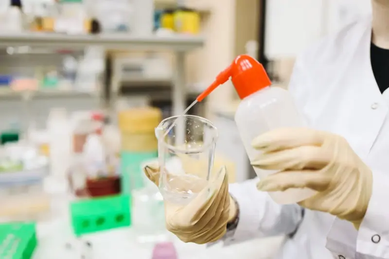 Gloved hands pouring clear liquid from a wash bottle into a beaker