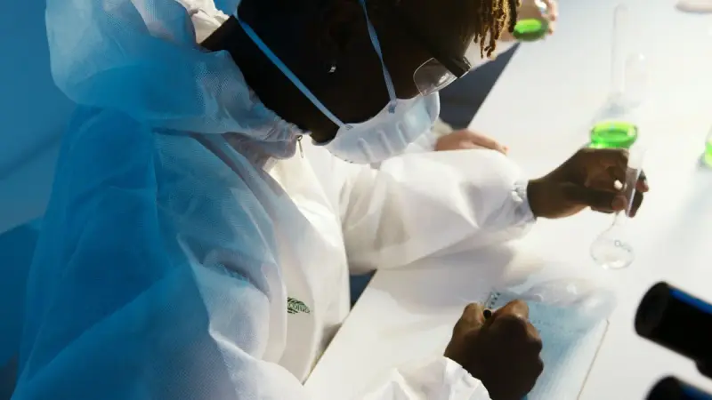 Researcher in protective gown and mask writing notes beside lab glassware