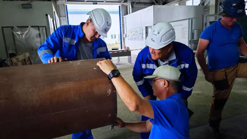 Four engineers in hard hats and blue workwear checking a large steel pipe section indoors
